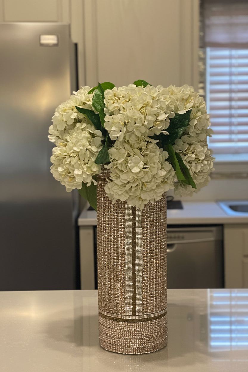 A tall, cylindrical vase covered in sparkling rhinestones, filled with a large bouquet of white hydrangeas with green leaves, set on a kitchen counter.