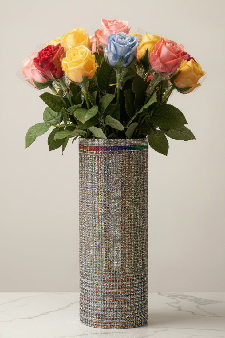 A tall cylindrical vase covered in iridescent crystal rhinestones, filled with a bouquet of multicolored roses displayed on a kitchen counter.