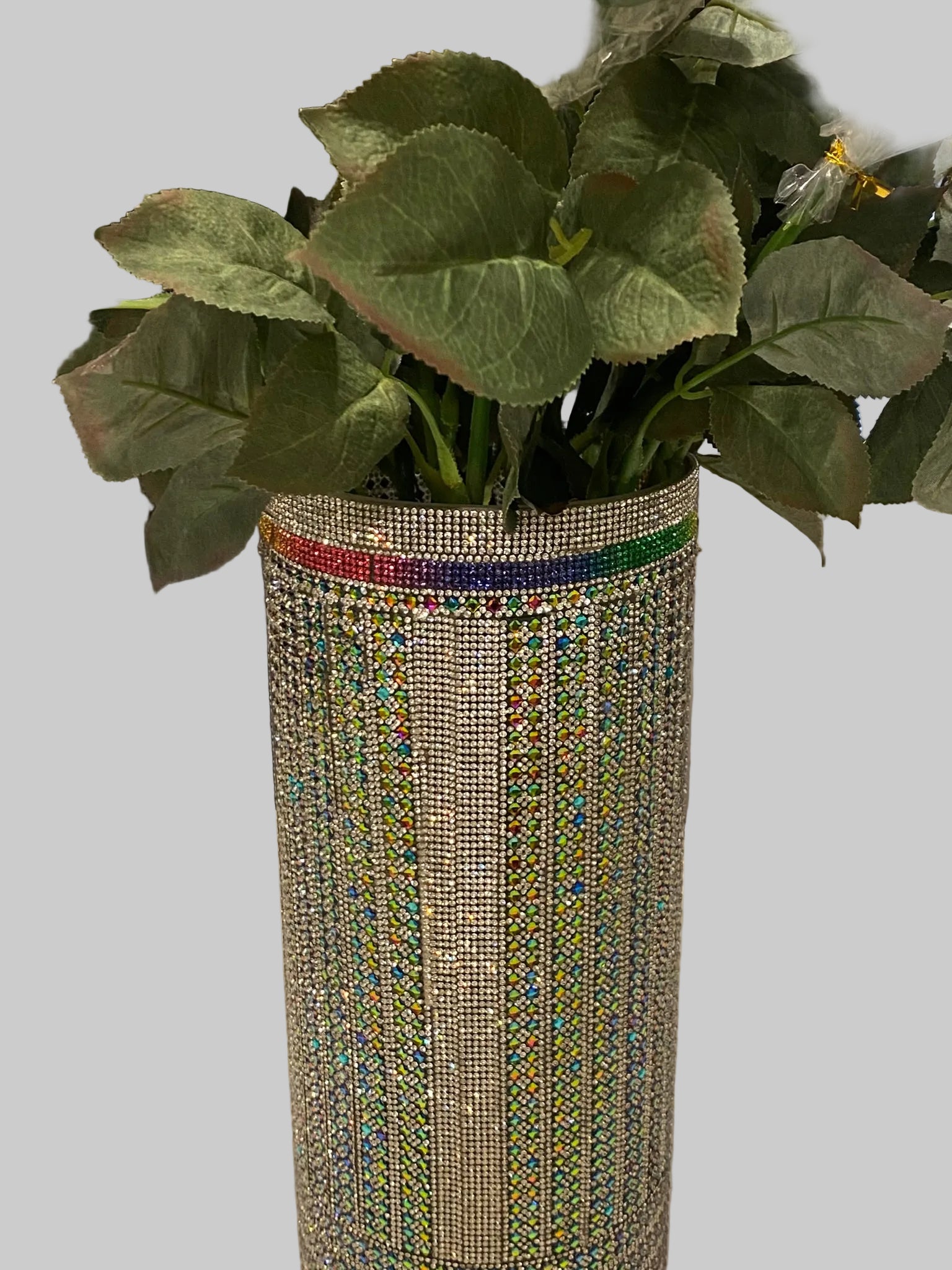 A tall 15" cylindrical vase covered in white crystal rhinestone with multi-color embellishments, holding a bouquet of green artificial-looking leaves.