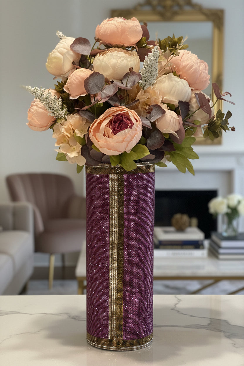 A tall cylindrical vase covered in sparkling purple crystal rhinestone gems with a vertical antique gold/silver stripe, holding a large faux floral arrangement of peach, cream, and mauve peonies with eucalyptus, green and gray leaves, set on a kitchen counter.